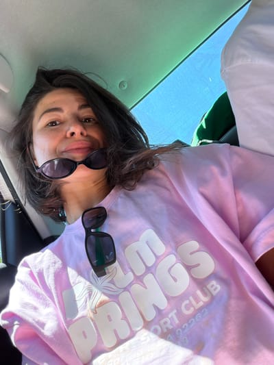The image captures a casual selfie of a person inside a car, taken on a bright, clear day in Kubasi, Montenegro. The photograph is oriented on its side, with the person's head to the left and their body extending to the right of the frame.
The central subject is a person with dark hair, looking directly at the camera with a neutral, slightly pouting expression. A pair of black sunglasses is worn pushed up on their face, and a second pair of black sunglasses hangs from the neckline of their shirt. The person is wearing a light pink, short-sleeved t-shirt.
Notable details on the t-shirt include large, white, rounded block letters spelling "SPRINGS". Above this, smaller white text reads "SPORT CLUB" followed by the number "02262". A white graphic, resembling a stylized starfish or a tropical leaf, is also visible on the shirt.
The setting appears to be a car interior. Above the person's head (which would be the ceiling if the image were rotated correctly), a light-colored car headliner is visible. To the right side of the frame, a clear, bright blue sky is visible through a window, indicating sunny weather during daytime. A glimpse of a dark green object, possibly a bag or another piece of clothing, can be seen behind the person's right shoulder. The activity is a self-portrait taken in a relaxed moment within the vehicle.