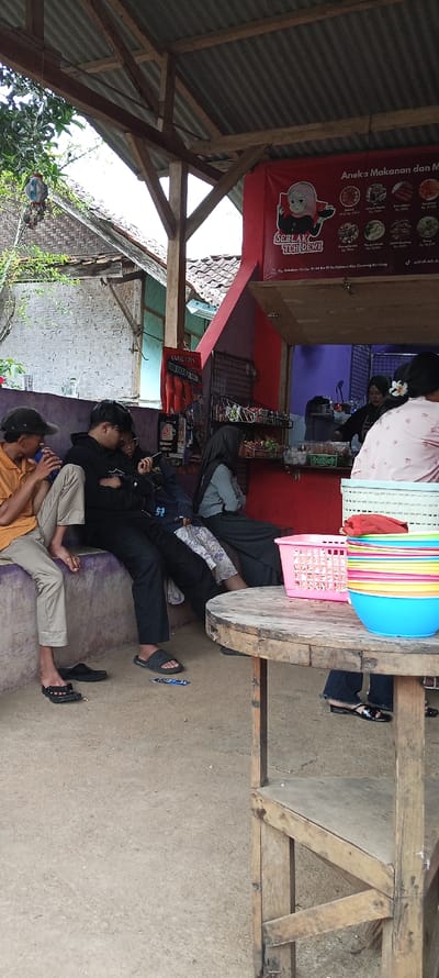 The image shows a scene in front of a street food stall in Cimaung, Indonesia (latitude: -7.074, longitude: 107.547). Four people are seated on a purple bench. The person on the left is wearing a yellow shirt and beige pants. The person in the middle is wearing a black shirt and black pants. The person on the right is wearing a blue hooded sweatshirt and a black skirt. A fourth person is wearing a black hijab and sitting with their back to the camera. There is a wooden table with several brightly colored plastic bowls and a pink plastic basket on top of it. There is a red sign behind the table, advertising food and drinks. The sign has a cartoon image of a woman wearing a red hijab. There is a building behind the sign. The scene is in front of a building with a corrugated metal roof and a wooden frame. There are several plants and trees in the background. The ground is a mixture of concrete and gravel.