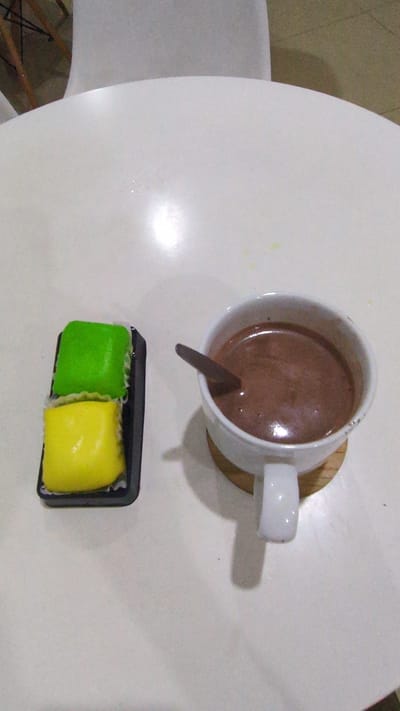 A top-down view shows a white table with a cup of chocolate drink, a spoon, and a small container holding green and yellow sweets. The cup sits on a wooden coaster. A white chair is visible in the lower left of the frame. The location is likely a cafe or similar establishment in Phan Rang – Tháp Chàm, Vietnam.