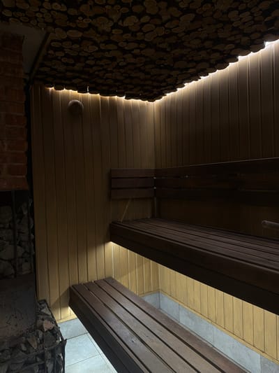 The image depicts the interior of a building, potentially a sauna or similar space, in what is stated to be Kamennoye, Russia. The walls appear to be constructed of horizontal wooden planks, with some areas darker and others lighter, suggesting different types of wood or finishes. One section of the wall is decorated with a dense arrangement of small, round wood pieces, possibly log slices, creating a textured effect. Adjacent to this, there is a brick structure, possibly a stove or fireplace base. In the lower right, there is a section of wall with a stone covering held in place by metal wiring. The lighting is dim, with a light source illuminating the edge of the log wall, accentuating its texture. Above, there are structural wooden beams. The overall atmosphere is rustic and dimly lit.
