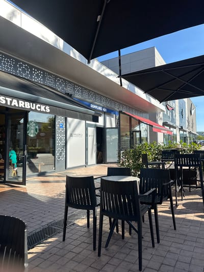 The image displays an outdoor paved plaza with seating in front of several retail storefronts at Mallorca Fashion Outlet in Marratxí, Spain, under a clear blue sky.
In the foreground, a light-colored rectangular paver stone plaza features a linear drainage grate on the left. Centrally, a round table with a light-colored surface is surrounded by four black outdoor chairs, each with slatted backs and armrests. To the right, additional black outdoor tables and chairs, including high-top configurations, are partially obscured by low, green ornamental bushes planted in rectangular beds. Strong, diagonal shadows from the left extend across the paving and seating area, indicating direct sunlight.
The midground consists of a row of storefronts. On the far left, a Starbucks coffee shop is visible, featuring large glass windows, a dark awning with "STARBUCKS" in white lettering, and the green siren logo on the glass. A person wearing a green top is partially visible inside the Starbucks near its open glass entrance door. Adjacent to Starbucks, two storefronts are labeled "MALLORCA FASHION OUTLET" with white signage on dark grey backgrounds; their glass facades are covered with white opaque material. One of these units has an open door, revealing a dimly lit interior with some visible wooden structures. Above these two units, a blue sign reads "DUMPLING HUB." Further to the right, a red awning and a sign for "HAWKERS" are visible. The building facade above the storefronts is light-colored, featuring a grey decorative panel with repeating geometric square and circular patterns. Large black cantilevered umbrellas extend over the seating area, providing shade.
The background reveals sections of a modern building with clean, light-colored paneling against the clear blue sky, and distant green foliage to the far right.