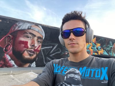 The image shows a selfie taken by a young man in Cartagena, Spain. He is wearing a dark t-shirt with a graphic of a motorcycle rider and the text "WORLDS BEST FMX MAGAZINE" and "MOTOX" in blue. He is also wearing large grey headphones and mirrored sunglasses. Behind him is a mural painted on a wall. The mural depicts a close-up of a man's face with a grey baseball cap, red paint markings on his face, and large hoop earrings. The background of the mural is black with abstract colorful shapes. The setting appears to be outdoors, with a blue sky and clouds visible. The ground in front of the mural is paved with concrete.
