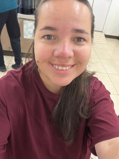 A close-up, selfie-style photograph features a young woman with light skin, blue eyes, and medium brown hair. She is smiling, revealing her teeth, and a mole is visible on her right cheek. She is wearing a maroon t-shirt. In the background, out of focus, a person is wearing a teal top and black pants. The environment appears to be indoors with tiled flooring and fluorescent lighting. The image does not provide specific geographical information to pinpoint a location within Chuo, Japan.
