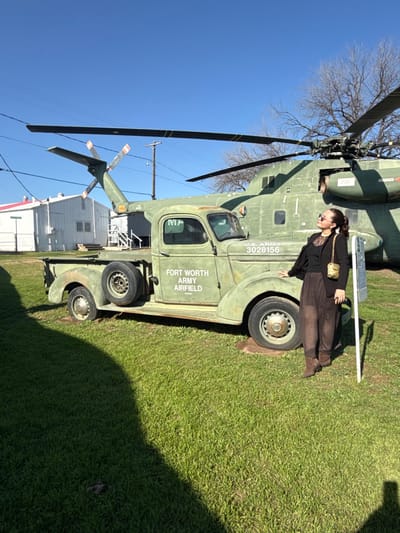 An army truck with “Fort Worth Army Airfield” painted on its side transports a green army helicopter. A woman in brown pants and a black top leans on the truck's front passenger-side tire. The helicopter has a number "3026156" printed on its side. The background is grassy and includes a building. The sky is clear and blue.