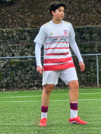 A young male soccer player stands on a green field. He wears a white jersey with red horizontal stripes and a team logo on the left breast. His shorts are white, and his socks are white with a red and blue band around the top. He's wearing red athletic shoes. Behind him are trees and a stone wall. A chain link fence runs along the top of the wall.
