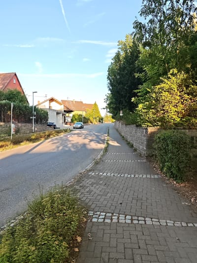 The image shows a street scene in Nordheim, Germany. The foreground features a paved sidewalk with a pattern of grey interlocking pavers and a decorative border of smaller cobblestones. To the left of the sidewalk is a narrow strip of green foliage. The sidewalk runs alongside a grey asphalt road.

On the right side of the road, a low stone wall runs parallel to the sidewalk, topped with dense green bushes and trees. Further down the road, on the left side, are several houses with red-tiled roofs and a modern white building. Two cars are visible on the road: a dark-colored car parked near the white building and a silver car driving away from the camera. The sky above is clear blue with a contrail from an aircraft and some wispy white clouds. The lighting suggests it is late afternoon or early evening. A speed limit sign with a "30" is visible on the right side of the road, near the stone wall.