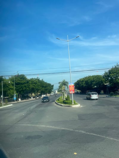 The image depicts a wide, paved road in Diên Lạc, Vietnam, under a clear blue sky with sparse white clouds. The road features multiple lanes, with clear white lane markings and some patches of darker asphalt. A central roundabout is visible, adorned with green shrubbery and palm trees. A tall, modern street lamp with multiple arms stands prominently in the roundabout.

Several vehicles are present on the road, including a black car in the foreground, a silver van in the middle ground, and a white truck further in the distance. A few other cars and motorcycles are also visible, suggesting moderate traffic.

Lining the sides of the road are lush green trees, some appearing to be mature and tall, while others are smaller, manicured bushes. Buildings are visible behind the trees on the right side of the road, with signs and awnings indicating commercial establishments. On the left side, there are more trees and some parked motorcycles, as well as a pole with a colorful banner displaying Vietnamese text and images. The overall environment appears to be a well-maintained urban or suburban area within Diên Lạc.