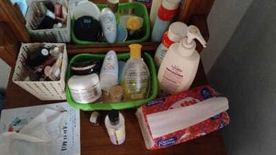 The image shows a wooden counter top with a collection of various hygiene and beauty products in containers. There are various plastic baskets containing the products. The products include:
- a white bottle with a pink lid and a white dispenser
- a yellow bottle with a white lid and a white label
- a white bottle with a blue lid
- a white bottle with a clear label
- a white bottle with a black label
- a clear container with a black lid
- a clear container with a white lid and a white label
- a container with a silver label with the word "WC" printed on it
- a white plastic basket containing a variety of lipsticks, makeup and brushes
- a white plastic basket containing a variety of cosmetic products including lipsticks, makeup, a clear container with a silver label and a small bottle of what appears to be hand lotion
- a green plastic basket containing a variety of cosmetic products including a clear container with a black lid and a small bottle of what appears to be hand lotion
- a clear plastic container with a white label and a black lid, containing a small bottle of what appears to be hand lotion
- a small, white, rectangular box with a blue label
- a white, rectangular box with a red label
- a white tissue on top of the red box.
The scene is located in a bathroom in the city of Panjalu, Indonesia, based on the latitude and longitude coordinates.
