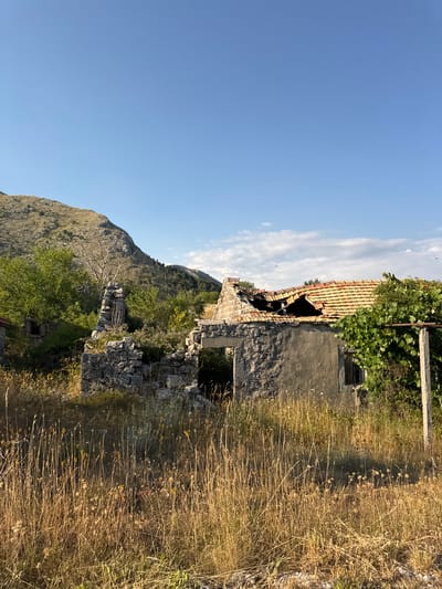 The image depicts a partially ruined stone house with a damaged roof, situated in the rural landscape of Kopito, Montenegro. The house is overgrown with vegetation, and the surrounding area is covered in dry grass and wild plants. In the background, a rocky, barren hill rises under a clear blue sky with a few scattered clouds. The scene suggests abandonment and the passage of time, with the natural environment reclaiming the man-made structure.