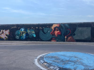 This image depicts a mural painted on a dark, textured wall, likely a seawall or retaining wall, bordering a paved roundabout. The mural features a large, stylized portrait of a man's head and shoulders, rendered in shades of red, orange, and teal. His right eye is open and appears to be looking forward, while his left eye is closed. A red square with a white "M" is superimposed on his cheek. To the left of the portrait are abstract graffiti tags in pink, yellow, and blue, with some illegible text. Further left, a blue and gold figure wearing a helmet and possibly a diving suit is depicted. To the right of the main portrait, there's a stylized red anchor shape and a smaller, grey rectangular panel with a stick-figure-like drawing of a person. The sky above is a pale blue with wispy clouds. The roundabout in the foreground is paved with asphalt and features a circular, raised island painted a vibrant, cracked blue with black and grey borders. The scene appears to be located outdoors in an urban environment, possibly near the coast, given the style of the mural and the presence of what could be a seawall. The location is identified as the "Paseo de Pereda" in the city of Cartagena, Spain, a prominent waterfront promenade.