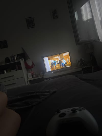 The image depicts an indoor scene, likely a bedroom, with a focus on a television screen displaying a group of people. The room is dimly lit, suggesting it is nighttime or the curtains are drawn. On the left side of the frame, a white dresser with drawers is visible, topped with various bottles and containers. A decorative gnome with a red hat and white beard stands on a shelf to the right of the dresser. The television is on a stand, and in the foreground, a white video game controller is placed on a dark surface, partially obscuring a person's leg and a grey blanket. In the background, a large window with white curtains is visible, and the wall displays two framed pictures. The scene appears to be set in a private residence, and there is no indication of it being located in Hausach, Germany.