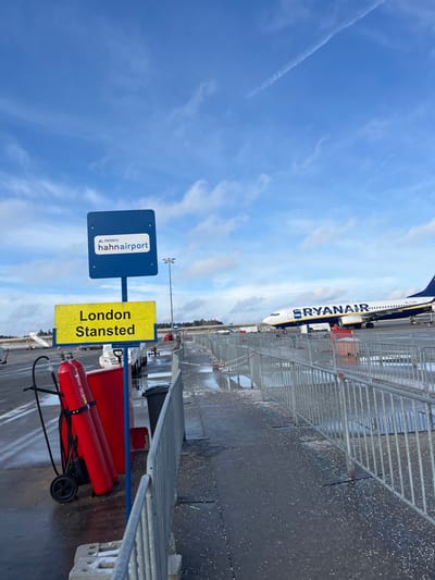 The image presents an outdoor airport scene at Hahn Airport in Lautzenhausen, Germany, under a partially cloudy blue sky featuring a prominent contrail. In the foreground and mid-ground, a designated walkway or queue area is established on a wet asphalt surface, scattered with white particulate matter, and bordered by silver metal crowd control barriers. To the left, a blue pole supports two signs: an upper blue sign bearing the "TRIWO hahnairport" logo and text, and a lower yellow sign indicating "London Stansted" in black capital letters. Situated near these signs are a black wheeled cart carrying two large red fire extinguishers and a black trash receptacle. In the mid-ground, a white passenger jet with "RYANAIR" branding in blue is stationary on the tarmac. Various pieces of indistinct ground support equipment are visible near the aircraft. No individuals are present or clearly identifiable in the scene.