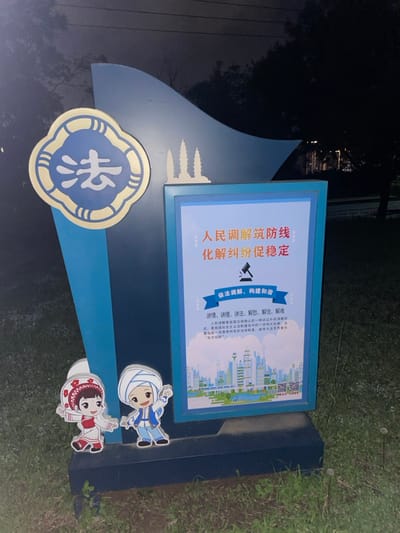 This nocturnal image captures a brightly illuminated outdoor display sign in Dali City, China, positioned on a grassy embankment with trees and dimly lit structures in the dark background. The sign, predominantly dark blue with a wavy top edge, features a circular gold-bordered emblem on its upper left, containing the white Chinese character "法" (fǎ, meaning "law"). To the right of this emblem, three white silhouettes resembling the iconic Three Pagodas of Chongsheng Temple are affixed.

A rectangular, internally lit panel dominates the right side of the sign. Its primary text in red reads: "人民调解筑防线 化解纠纷促稳定" (People's mediation builds a defense line, resolves disputes, promotes stability). Below this, a blue banner states: "依法调解、构建和谐" (Mediate according to law, build harmony), accompanied by a gavel icon. Further text details mediation principles, and a cityscape illustration with buildings, wind turbines, and a QR code occupies the bottom portion of the panel.

At the base of the sign, two cartoon character cutouts are present. The left character is a girl in red and white traditional attire, while the right character is a boy in blue and white traditional attire with a white head covering, both depicted with arms outstretched. The scene is static, devoid of human activity, and illuminated solely by the sign's internal lighting.