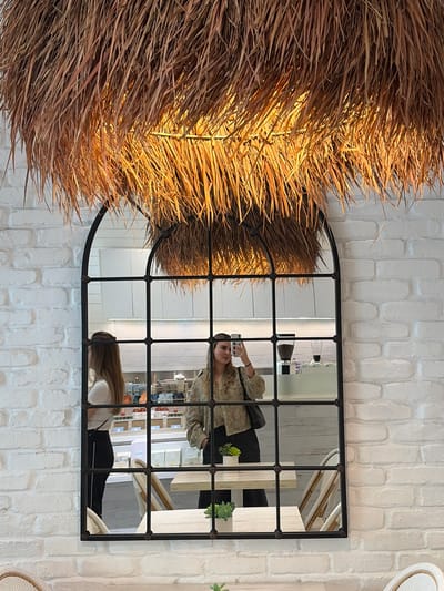 A portrait-oriented photograph shows a decorative display in a shop interior. A large, black-framed arched mirror is mounted on a textured white brick wall. The mirror reflects the interior of the shop, including shelves stocked with products and two figures. One figure is holding up a cell phone, presumably taking a picture, and the other is bending over or looking at something on a lower surface. To the left of the mirror is a large decorative display made of dried straw or reeds that partially obscures the mirror. The setting appears to be in a retail space, likely in Coral Gables, United States of America.