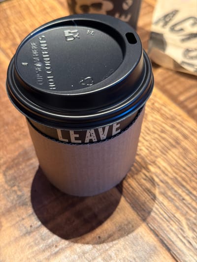 The image displays a disposable coffee cup with a black plastic lid and a corrugated cardboard sleeve, positioned on a light brown wooden table. The black lid features an oblong drinking opening, a raised rim, and embossed text stating "CUP SEAM HERE" and "HOT CONTENTS," alongside a partial recycling symbol and a utensil icon. The natural brown cardboard sleeve displays the word "LEAVE" in white, stippled capital letters against a dark background band. The wooden table surface exhibits distinct grain patterns, suggesting horizontal planks. Strong directional lighting from the upper left casts a clear, elongated shadow of the cup onto the table. In the blurred background, a second, dark-colored disposable cup with light patterns is partially visible, along with a crumpled brown paper item displaying indistinct light text. No individuals or specific actions are depicted. The scene is consistent with a commercial establishment such as a coffee shop or cafe located in Bretton, United Kingdom; however, a precise address within Bretton cannot be determined from the visual evidence.