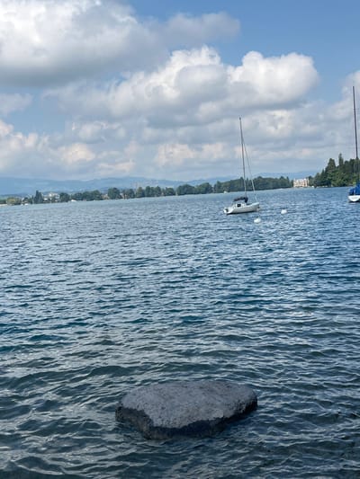 The scene depicts Lake Thun, viewed from Hünibach, Switzerland. In the lower-center foreground, a dark grey, irregularly shaped rock is partially submerged in the rippled, dark blue water. Two sailboats are present in the midground water. A white-hulled sailboat with a visible mast and furled sails is anchored slightly right of center, accompanied by two white buoys near its hull. A second sailboat, partially visible on the far right, features a blue covering over its bow and a visible mast. The distant background comprises a tree-lined shoreline, interspersed with structures, and low-lying mountains visible on the horizon. The sky is characterized by a blue expanse with numerous scattered white cumulus clouds. No individuals are visible in the image.