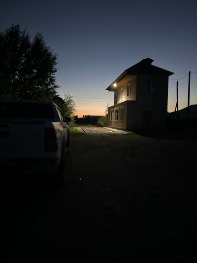 A white pickup truck is parked on a dirt road facing away from the camera on the left side of the frame. To the right, a two-story building with a pitched roof is illuminated by a single light fixture on the upper facade. The building appears to be constructed of light-colored brick or siding. A metal staircase is visible on the left side of the building. In the background, a dark silhouette of trees is present on the left, and a bright orange and yellow sunset is visible on the horizon. Telephone poles and wires are also visible in the distance. The scene is set at dusk, with the sky transitioning from a deep blue to a lighter hue near the horizon.