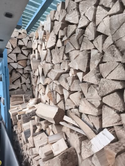 A large stack of cut firewood is stored under a corrugated metal roof structure, partially enclosed by a blue metal frame on the left. The firewood is neatly piled, with individual logs split and stacked in an organized manner. The scene appears to be an outdoor storage area, possibly a wood shed or a covered area adjacent to a building. The lighting suggests it is daytime, with natural light filtering through the roof. The image does not provide any specific visual cues to pinpoint its exact location within Eitorf, Germany, but it depicts a common scene of a wood supply for heating.