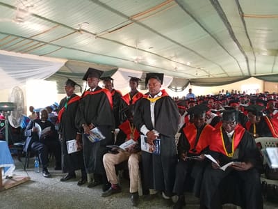 The image shows a graduation ceremony taking place inside a large hall in Rumde Waziri, Nigeria. The hall is decorated with white drapes and has a green tiled roof. There is a stage at the front of the hall where the graduates are sitting. The graduates are wearing black gowns with red sashes and black caps. Some of them are holding certificates. The graduates are seated on plastic chairs with their faces turned toward the front. At the back of the hall, a crowd of people are sitting and watching the graduation ceremony. There are many large ceiling fans in the hall. There is a large metal fan in the left foreground.