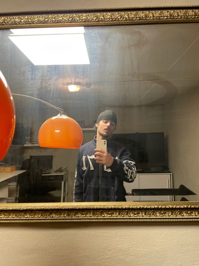 A male individual is depicted in a mirror selfie taken within an indoor setting in Oberhausen, Germany. The subject, a male with facial stubble/beard, wears a dark beanie and a dark blue zippered hooded sweatshirt featuring white or light-colored text/graphics on the chest (appearing as "TMF" or similar) and stylized text on both sleeves. He holds a light-colored smartphone (likely an iPhone) in his right hand, positioned to capture his reflection.

The mirror itself is large and rectangular, framed by an ornate, gilded molding, and exhibits some surface smudges or dust. Reflected in the mirror, the background reveals a room with light-colored walls. Dominating the mid-left foreground of the reflection is an orange, dome-shaped lamp with a curved silver arm, partially obscuring the view. To the left background of the reflection, a desk with a dark computer monitor is visible. To the right background, a white low-profile storage unit or cabinet is present, with a dark, angled object (possibly furniture) partially visible in front of it. Overhead lighting reflected in the mirror includes a bright, rectangular ceiling panel and a smaller, yellowish-orange industrial-style fixture. The man stands facing the mirror, engaging in the act of photography.