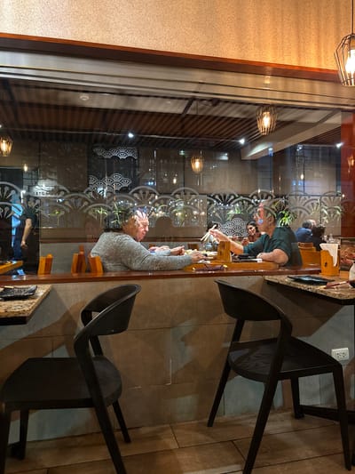 The image shows an interior view of a restaurant in Magdalena del Mar, Peru, likely a sushi restaurant, as indicated by the decorative elements suggestive of Japanese art and the presence of chopsticks and dishes. The scene is viewed through a large window, with reflections overlaying the interior details. In the foreground, two modern black chairs are positioned at tables.
The main subjects are two individuals seated at a counter, viewed from the side through the glass. The person on the left, appearing to be an older woman with gray hair, is wearing a gray sweater and is looking down at something on the counter, with her hands near a plate of food. The person on the right, also an older woman with dark hair, is wearing a dark green top. She is holding chopsticks and appears to be interacting with food on her plate, looking towards the other person. There are other patrons visible in the background, some blurred and less distinct, suggesting a bustling atmosphere.
The restaurant interior features wooden accents, reflective surfaces, and decorative elements with a fan-like motif. Lighting fixtures are visible, including pendant lights that cast a warm glow. The overall impression is of a well-lit dining establishment. The reflections on the window show the exterior environment, though it is difficult to discern specific details due to the blur and the light conditions.