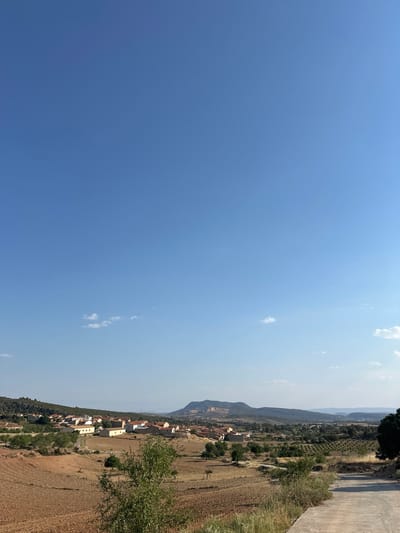 The image captures a panoramic view of a rural landscape under a clear blue sky with scattered white clouds. In the mid-ground, a village with red-tiled roofs and white-washed buildings is nestled amongst rolling hills. The foreground features dry, brown fields, possibly recently plowed, with sparse green vegetation and trees. A concrete path or road curves from the lower right corner into the scene. In the background, a mountain range with eroded features is visible, suggesting a semi-arid environment. The overall impression is one of a tranquil, sun-drenched rural setting in the municipality of Alpuente, Spain. There are no people or animals visible in the image.
