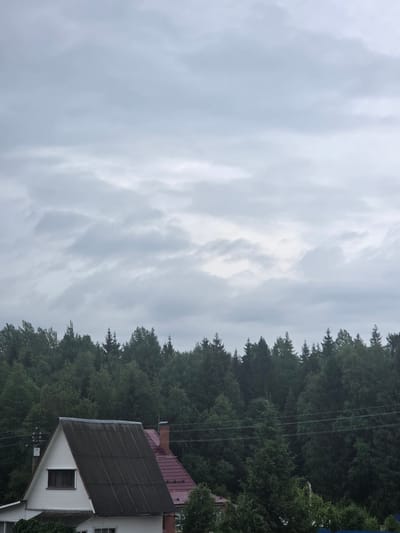 The image depicts a cloudy sky and a forested landscape with a house in the foreground. The sky is overcast with soft, diffused light, suggesting it might be daytime, possibly late afternoon or early morning. The trees are dense and green, forming a natural border to the scene, with some appearing to be coniferous. A portion of a white house with a dark, corrugated roof and a red tiled roof is visible on the right side of the image. A single window with dark trim is present on the white facade of the house. Power lines can be seen running through the trees, indicating the presence of human infrastructure. The overall mood is serene and perhaps a little somber due to the cloudy weather. The location is identified as Восход Снт (фролово), Russia. There is no text visible in the image.