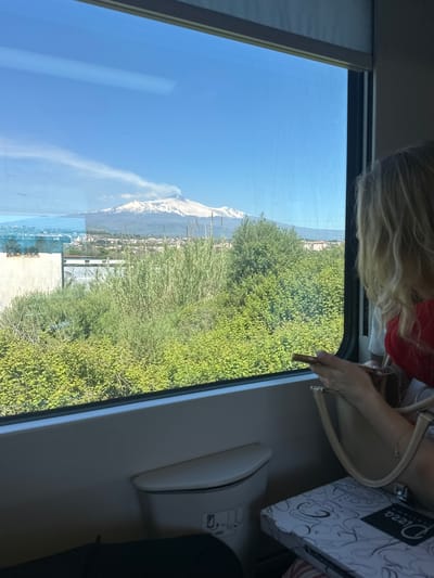 Here is a forensic description of the image:

The photograph, taken from inside a moving vehicle, features a scenic view of the urban area near Acireale, Italy. The background showcases Mount Etna, its peak partially covered in snow, under a clear blue sky. Lush green vegetation fills the midground, obscuring some of the urban structures.

A blonde-haired person wearing a red top is partially visible at the top of the frame. They are holding a light-colored purse. A "Pizza" branded box sits on the vehicle's interior surface. The box has a white and black design. A car seat and a glimpse of black clothing can also be seen within the vehicle's interior.