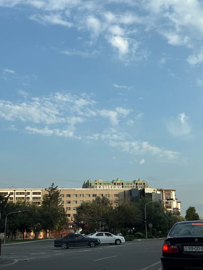 This is an eye-level, slightly low-angle, medium shot of a street scene in Baku, Azerbaijan, with a focus on buildings and vehicles. The sky is a clear blue with scattered white clouds. Dominating the background is a multi-story building with a prominent "SAMSUNG" logo on its upper facade, and another sign reading "SEVEN SPACE". The architecture of this building features a stepped design with a green roof. To the left of this building, another building with numerous windows is visible. In the foreground, a black car with an Azerbaijani license plate (99-GO-8-) is stopped at what appears to be an intersection. A white car is also visible in the middle distance, and various trees line the street. The overall scene suggests an urban environment during daylight.