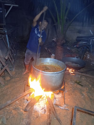A man is stirring a large pot of food over a fire in Keudeteupinpunti, Indonesia. The man is wearing a blue t-shirt and shorts, and he is barefoot. The pot is made of metal and is filled with a brown, stew-like substance. The fire is made of wood and is burning brightly. In the background, there is a palm tree and some debris. The ground is dirt.