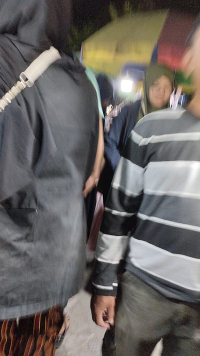 In the city of Sukodono, Indonesia, a group of people are visible in what appears to be an outdoor gathering, possibly a festival or market. The image is blurry due to motion. A person in the foreground on the left is wearing a dark, loose-fitting garment, with a pale strap across their shoulder. To their right, another individual is wearing a dark shirt, and behind them, a person in a hijab with glasses is looking to the right. The central figure in the foreground is wearing a striped long-sleeved shirt with black and white horizontal bands and dark pants. The background shows a blurred yellow structure, suggesting tents or stalls, and a bright light source, possibly a lamp. The ground appears to be paved or dusty. The overall impression is a busy, dimly lit environment with people in motion.