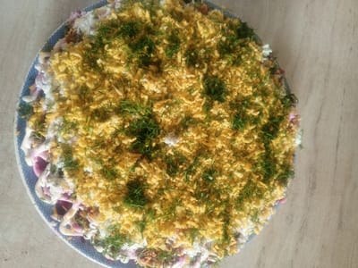 A top-down, slightly angled medium shot captures a layered salad presented on a round, blue and white patterned plate. The salad is generously topped with finely grated yellow egg yolk and sprinkled with chopped fresh dill, creating a textured, vibrant surface. Beneath this topping, layers of white ingredients, likely egg whites and mayonnaise, are visible, interspersed with glimpses of reddish-purple elements, possibly beetroot. The salad appears to be freshly prepared and is placed on a light-colored, textured surface, likely a countertop or table. The lighting suggests it might be daytime, but there are no specific cues for time of day, weather, or a distinct geographical location beyond the general context of Buraly, Russia. No people or text are visible in the image.