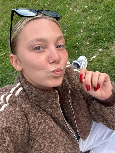 The image shows a young woman lying on a patch of green grass. Her face is angled towards the camera, and she has a nose ring and is puckering her lips. Sunglasses are placed near her head on the grass. She is wearing a brown jacket with white stripes, and her nails are painted red. In the background, a sneaker is visible. There is no information to confirm that the location is Marquette-lez-Lille, France.