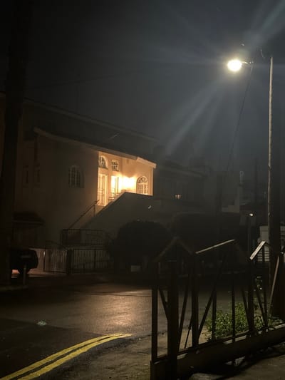 The image depicts an outdoor night scene in Egkomi Lefkosias, Cyprus. A bright street light, mounted on a utility pole on the left, is the primary light source, casting strong rays across the dark sky and illuminating the immediate foreground. To the right, a multi-story building with a light-colored facade is visible, featuring several windows, some of which are distinctly arched, glowing with warm yellow light from within.

The ground, appearing to be an asphalt road or pathway, is wet and reflective, suggesting recent rain. A prominent yellow double line runs along the right side of the wet ground. A dark, metal fence or gate structure stands between the building and the road, partially obscuring some dark foliage behind it. Near the bottom right, a dark, rectangular object, possibly a bin or equipment, is situated on the ground.

The time of day is clearly night, characterized by the deep darkness and the reliance on artificial lighting. The wet ground adds a notable detail to the scene, reflecting light and creating a slightly moody atmosphere. No people are visible, and no specific activity is taking place; the scene is static. On a white surface or box near the base of the building, some faded or indistinct text is visible, but it is illegible due to distance and lighting.