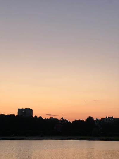 The image depicts a serene sunset scene over a body of water, likely a river or lake, in Salihorsk, Belarus. The sky transitions from a pale lavender at the top to a warm orange hue near the horizon. Silhouetted against the vibrant sky is a dark tree line that runs along the water's edge. Beyond the trees, several buildings are visible. On the left, a multi-story, block-like residential building stands prominently. To the right of this, a red-domed orthodox church with a cross on top is partially visible, nestled within the trees. Further to the right, more apartment buildings with illuminated windows can be seen. The water in the foreground reflects the colors of the sunset, creating a calm and tranquil atmosphere. The overall impression is one of peaceful evening settling over the city.