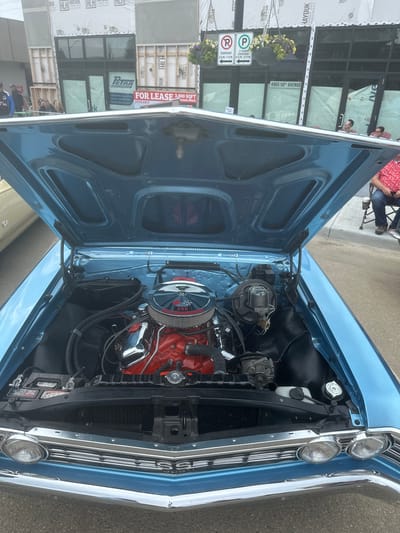 The image shows the open hood of a light blue classic car, revealing its engine. The car is parked on a street in front of a building undergoing construction. In the background, there is a sign that reads "FOR LEASE 3,000 SQFT LEASE SPACE," indicating a commercial property. Another sign on the building shows the address "4905 50th Avenue." This address corresponds to a location in the City of Leduc, Alberta, Canada. There are several people visible in the background, some standing and others sitting in chairs, suggesting an outdoor event or gathering. The overall scene depicts a street view with a focus on the vintage automobile and its engine.