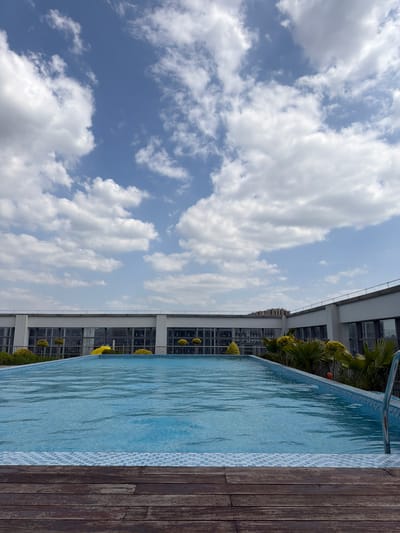 The image captures a rooftop swimming pool adjacent to a modern building under a partly cloudy sky, likely in the Linping District, China. The pool, filled with clear blue water, is surrounded by wooden decking. The building is a multi-story structure with glass windows. The sky is blue with scattered white clouds. The photograph is oriented in a portrait format.