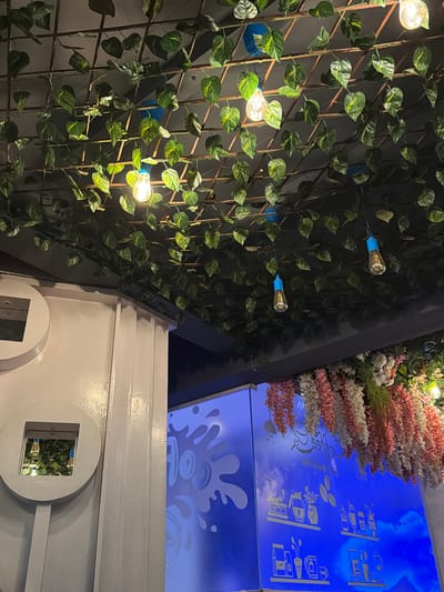 The image captures the interior of a cafe, likely in Shamshabad, India, with a focus on the ceiling and upper walls. The ceiling is adorned with a network of artificial green vines interwoven with a metal grid. Several filament light bulbs are suspended from the ceiling, some with decorative blue accents.
On the left side of the image, a white pillar structure features a circular design with a square opening, revealing a reflection of the ceiling and a light fixture within. Adjacent to this, a blue illuminated wall displays etched graphics related to coffee and tea, including images of coffee beans, grinders, kettles, and cups. Cascading floral arrangements in shades of pink, white, and green hang from the ceiling above this wall. The overall ambiance suggests a decorative and themed cafe environment. No people are visible in the frame.