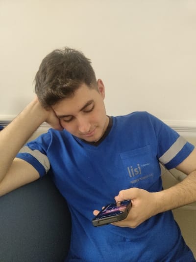 A young man, wearing a blue t-shirt with a white reflective stripe on the left sleeve and a white logo on the right chest that reads "lisi," sits in a chair and uses his right hand to hold a black smartphone in front of him. His left arm is resting on his left leg, and his left hand is propped on his head. The man's hair is short and dark, and he appears to be looking down at his phone. The background of the image is a white wall. The scene appears to be an interior domestic setting, though the location is not precisely clear. The image is consistent with a location in the city of Izmir, Türkiye, at the provided coordinates (latitude: 38.332, longitude: 27.124), a residential area known for its contemporary housing.