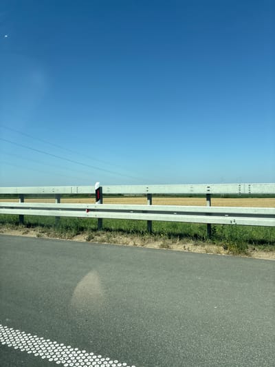 A wide-angle, eye-level, outdoor shot captures a rural roadside scene under a clear blue sky in Ruma, Serbia. In the foreground, a portion of an asphalt road with a distinct white circular marking is visible on the lower left. Beyond the road, a green grassy verge transitions to a metal guardrail. The guardrail, composed of two horizontal silver bars supported by vertical posts, runs across the middle ground of the image. Behind the guardrail, a field of dry, golden crops stretches to the horizon. In the distance, a hazy landscape features power lines and electricity pylons, indicative of rural infrastructure. The overall impression is one of a sunny day on a rural road, with agricultural land and utility structures present in the environment. There are no people or animals visible in the frame.