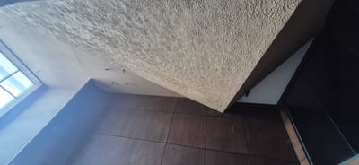 This media file captures an interior architectural detail, likely within a modern building in Ashgabat, Turkmenistan. The perspective is from below, looking upwards at a textured, beige ceiling that slopes dramatically. The texture of the ceiling appears rough and uneven, possibly made of concrete or a similar material with a textured finish.
Adjacent to this textured ceiling is a smooth, darker brown surface, likely a wall or another architectural element, divided into large square panels. The lighting suggests it is daytime, with natural light streaming in from a large window or skylight system on the left side of the frame. The window has multiple panes, and the light appears bright, indicating a sunny day.
Several small, dark objects, possibly light fixtures or mounting hardware, are visible against the smooth, lighter-colored ceiling near the textured section. The scene is devoid of people or any overt activity. The overall impression is one of clean, modern lines and varied textures within an interior space, possibly a lobby, atrium, or a large public area. The dark, glossy surface on the right suggests a reflective material, possibly glass or polished stone, contrasting with the rough ceiling and paneled wall. There is no visible text in the image.