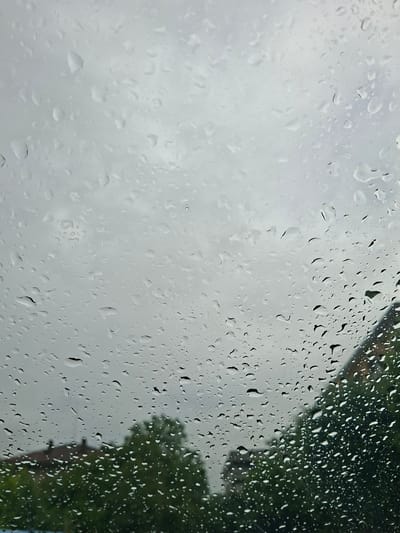 The image shows a rain-streaked window. Outside, on the left, is a blurry view of overcast, gray sky. On the right, also out of focus, are green trees and a blurred building outline, possibly a house. The location is Galabovo, Bulgaria. There are no people visible. The scene conveys a rainy day.