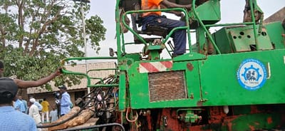 The image captures an outdoor scene featuring a large, weathered green heavy machinery, likely a piece of construction or road-building equipment, under what appears to be an overcast or diffused daylight sky.

In the foreground and central part of the image, the prominent feature is the robust green machine. It shows signs of wear, including rust spots on its metal panels, and features red and white striped caution decals on its side. Various mechanical components, including hydraulic lines and grills, are visible. An operator, clad in an orange safety vest and dark blue work trousers, is seated in the elevated control cab of the machine, his back facing the viewer, as he operates the controls with his right hand.

To the left of the machine, a man with dark skin, wearing a dark cap and a light blue shirt, is holding onto a green metal rail that forms part of the machinery's structure, his left arm extended. Below his arm and at the base of the machine, several cut logs or pieces of wood are visible, suggesting either material being processed or debris from an ongoing work.

In the mid-ground, behind the man holding the rail, a group of people is gathered. Several individuals are wearing light blue shirts, with one person in the center holding what appears to be a microphone or a small camera, indicating possible media presence or reporting on the event. A person in a yellow shirt is also visible further back within this group, and a child can be faintly seen near the ground.

The background consists of lush green foliage from a large tree, whose bare branches are also visible, contrasting with the machinery. A building with a brown roof and lighter-colored walls can be seen behind the tree and the gathering of people, suggesting a semi-urban or construction site setting.

A blue and white circular logo is affixed to the right side of the green machine. The text on the logo reads:
- "PRESENSIO CONSTRUCTION LTD." (The first part "PRESENSIO" is partially clear but discernible.)
- Below the company name, there is an emblem depicting a gear, a stylized cityscape with buildings, and possibly an abstract figure.
- Below the emblem: "RC: 78107193"
- And beneath that: "7807193" (possibly a registration number or an abbreviated phone number).

The overall impression is one of an active construction or work site, possibly involving a public event or media coverage, focused on the operation or display of the heavy machinery.