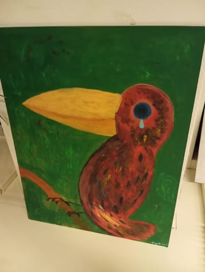 The image displays a painting of a red bird with a large yellow beak and a blue eye with a teardrop. The bird is perched on a brown branch against a green background. The painting is signed "oboziq" in the bottom right corner. The painting is displayed on a wall indoors, with a white and cream-colored surface visible in the foreground and background. The location in Anderlecht, Belgium, is not discernible from the image itself.