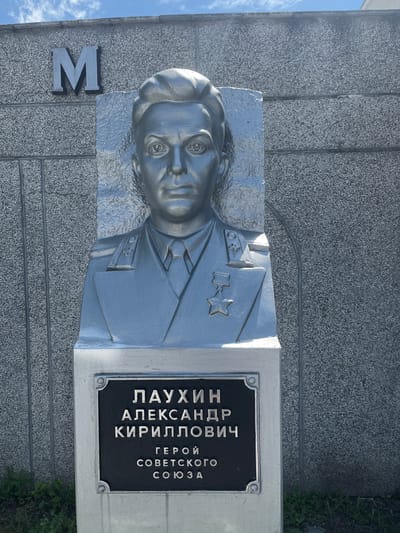 The image shows a silver-colored bust of a man in military uniform, mounted on a pedestal. The bust is positioned in front of a textured gray stone wall. Above and to the left of the bust, the letter "M" is visible, also appearing to be made of metal. The pedestal has a black plaque with text inscribed in silver lettering. The text on the plaque reads: "ЛАУХИН АЛЕКСАНДР КИРИЛЛОВИЧ" and below that, "ГЕРОЙ СОВЕТСКОГО СОЮЗА". This translates to "LAUKHIN ALEXANDER Kirillovich" and "HERO OF THE SOVIET UNION". Green foliage is visible at the base of the pedestal. The sky is visible at the top of the image, with some clouds. Based on the inscription on the plaque, this memorial is dedicated to Alexander Kirillovich Laukhin, a Hero of the Soviet Union. It is located in the village of Takhtamukai, Takhtamukaysky District, Republic of Adygea, Russia, at the monument to Heroes of the Soviet Union.