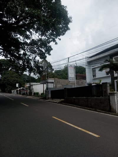 The image captures an outdoor street scene under an overcast sky in the city of Ariyalai East, Sri Lanka. A two-lane asphalt road, marked by a single dashed yellow line down its center, dominates the lower half of the frame, receding into the distance.
On the right side of the road, a grey concrete block wall topped with a dark fence encloses a property. A decorative white pillar marks an entrance. Behind this, a modern, multi-story white building with dark window frames is visible. Adjacent to the building, a tall white sign with red and black text reads "PRAKTIK DOKTER BERSAMA" (Together Doctor Practice) and lists services and operating hours, also displaying the name "CIDAHU". Numerous black electrical or communication wires traverse the upper mid-ground, strung between utility poles. Beyond the primary white building, older, less defined structures and dense green foliage on an elevated terrain are present.
On the left side of the road, a broad canopy of dark green tree leaves occupies the upper left quadrant. Below this, a paved shoulder or narrow sidewalk runs parallel to the road. Further down the street, a series of more modest, mixed-use structures are visible, one featuring a red roof. A dark-colored scooter or motorcycle is parked near one of these structures, and a single, indistinct individual is standing in its vicinity. The scene indicates an urban or semi-urban environment with noticeable tree coverage.