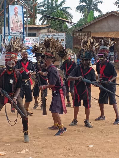 The image depicts a group of approximately seven men engaged in what appears to be a cultural or ceremonial event in Omicha Umu Awada, Nigeria, under bright daylight. The men are dressed in traditional attire, consisting of black shirts and shorts or trousers, adorned with striking red sashes or strips of fabric crisscrossing their chests and waists. Many wear large, elaborate feathered headpieces, and some have facial markings, such as white paint on their chins and around their eyes, or yellow/green adornments in their mouths.
Several participants are holding long, rifle-like objects, which could be traditional firearms or ceremonial staffs. Their stances suggest they are in motion, possibly performing a dance or participating in a procession. Some men wear ankle adornments, and their footwear includes modern sneakers.
In the background, unpaved ground gives way to buildings with corrugated iron roofs, surrounded by lush green foliage and palm trees, indicating a rural or semi-urban setting.
Various signs are visible in the background:
* A large banner features a portrait of "CHIEF AA CITERAS."
* Another sign partially reads "THE LIC OF YAHWEH SYNAGOGUE INCORPORATED."
* A sign advertises "FREE MEDICAL OUTREACH."
* A directional sign clearly states "THIS WAY TO EBEREMPU BOTORCOS COMMUNITY, EZINIHITT MBA LOCAL GOVERNMENT AREA," providing specific location details.
* A building in the background is identified as "EZUDO GIRLS SCHOOL," with "EZUDO EZI" partially visible below it.
The scene captures a vibrant community gathering, showcasing traditional customs and attire in a communal setting.