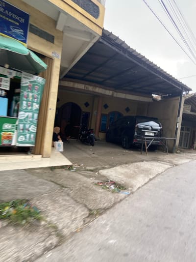 The scene is a residential street in Kecamatan Margaasih, Indonesia (latitude: -6.965, longitude: 107.541). A black SUV is parked under a carport attached to a residential building, with the license plate number "D 1604 AJ". A motorcycle is parked in front of the car, and there is a person sitting on the ground near the vehicle. There are various objects scattered on the street, including a small white box and some garbage. The buildings are all yellow with metal roofs. There is a sign with the word "GRAFISOI" on it and a green awning hanging from the building on the left. The buildings are decorated with diamond-shaped tiles and a faded blue door. The street is paved with concrete, and the sidewalk is made of cement. There is a green patch of grass on the side of the street. The weather appears overcast, with a few wires visible overhead.
