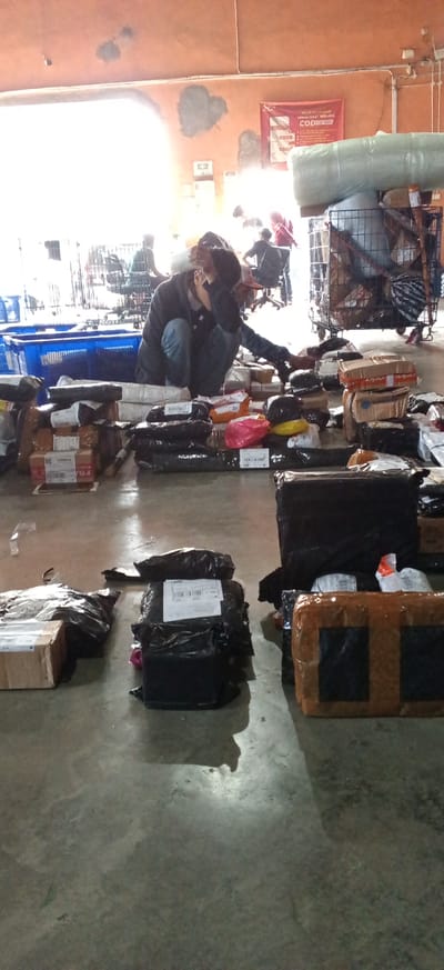 The image depicts a warehouse or logistics facility, likely located in Kiangroke, Indonesia. The scene is filled with numerous packages and parcels of various sizes, indicating an active shipping and receiving operation. In the foreground and midground, stacks of brown cardboard boxes and black plastic-wrapped items are scattered across the concrete floor.

A person, dressed in a dark hooded jacket and blue jeans, is crouched down among the packages, appearing to be sorting or organizing them. Their face is partially obscured, but they are touching their head with one hand, suggesting a moment of concentration or fatigue. In the background, other individuals are visible, some seated on chairs and others standing, also surrounded by boxes. One person is pushing a cart filled with items.

The environment appears to be indoors, with a textured, orange-colored wall in the background. Overhead lighting is present, though some areas are bright and potentially overexposed. A large, light-colored cylindrical object, possibly wrapped in plastic, is visible on a metal cart on the right side of the image. A red banner with text, likely a company logo or advertisement, is also mounted on the wall in the background. The overall impression is one of a busy, functional workspace dedicated to package handling.
