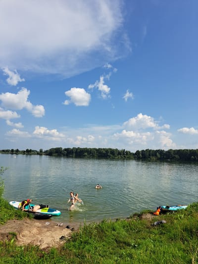 In Sorochii Log, Russia, on a sunny day with scattered white clouds against a blue sky, a group of individuals are engaged in water activities by a lake. In the foreground, on the grassy bank, are two paddleboards. One paddleboard has a person lying on it wearing a life vest and swim trunks, and another person sitting on the edge with a life vest. Near them, a black inflatable ring is partially submerged in the water. Further into the lake, two children are splashing and playing, with one child appearing to jump. Another person is visible in the distance, swimming. On the right side of the bank, a blue kayak with orange life vests is present. The background features a dense line of green trees across the lake, suggesting a natural landscape. The water is calm with ripples.